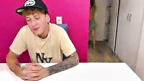 Tattooed guy in cap and NKY shirt sits at table with pink wall and blurred woman in background