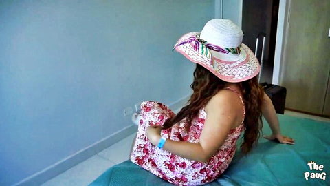 Sexy Latina babe in pink floral dress and hat sitting on bed showing curves from behind