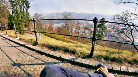 Person's legs in dark pants and shoe resting by black metal railing overlooking scenic cityscape and hills