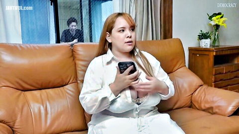 BBW East Asian wife with huge tits exposes deep cleavage holding phone on couch as man spies behind glass