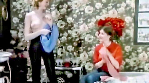 Topless blonde with bare perky tits holding blue scarf smiles at seated brunette in vintage floral bedroom