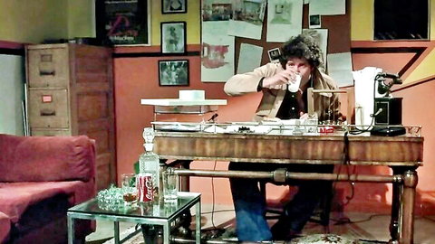 Vintage 1978 adult film scene: curly-haired man drinking in office