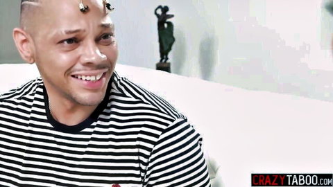 Bald mixed-race man with forehead piercings smiling in striped shirt on couch