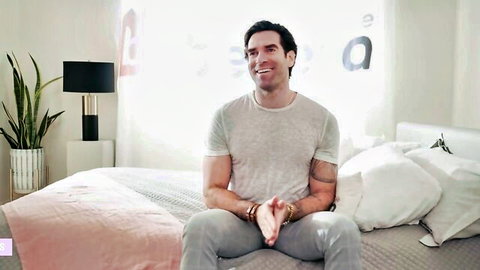 Handsome smiling stud in gray shirt sits on pink bed during flirty bedroom interview