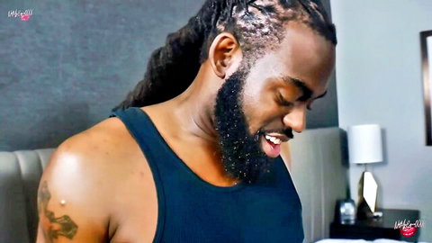 Ebony stud with dreadlocks and beard smiling seductively in tank top on bedroom bed
