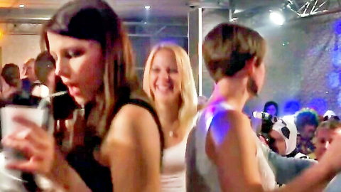 Blurry party scene with smiling brunette and blonde women holding drinks, teasing at club orgy