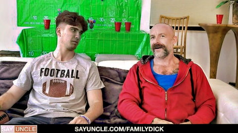 Bald older man and young football fan nephew cozy on couch in gay scene