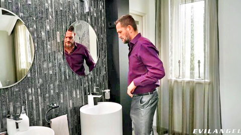 Mature man in purple shirt stands in bathroom mirror adjusting pants crotch area
