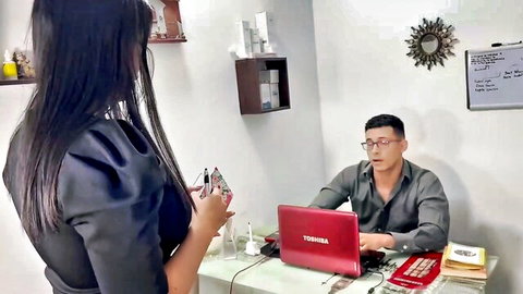 Brunette secretary in tight blouse stands over Asian boss typing on red laptop in office