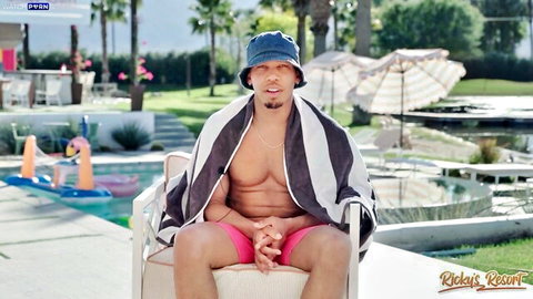 Shirtless muscular guy in bucket hat pink trunks and towel sits poolside at Riley Reid resort
