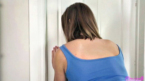 Brunette MILF in tight blue tank top peeking around white door from behind