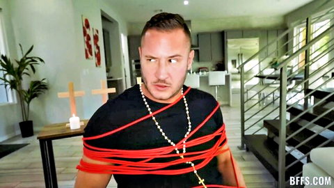 Bound shirtless man with red ropes and cross necklaces in intense BDSM living room setup