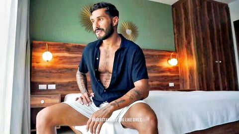 Bearded muscular man in open blue shirt and white shorts, teasing pose on bed