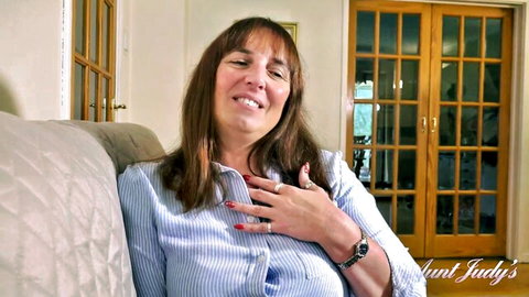 MILF Aunt Judy smiling with hand on chest in blue blouse on couch