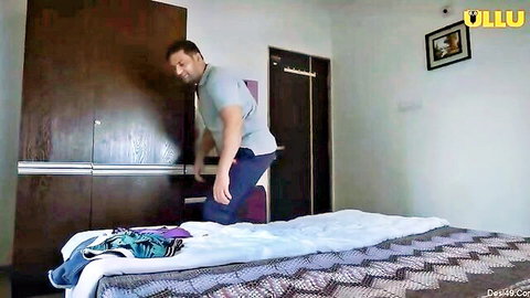 Indian man in shirt and pants leaning on wooden wardrobe in bedroom with colorful clothes on bed