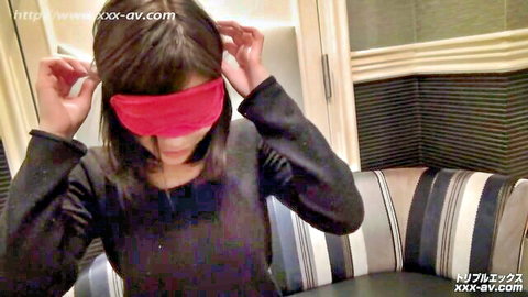 Blindfolded Asian woman in black top adjusting pink blindfold on couch