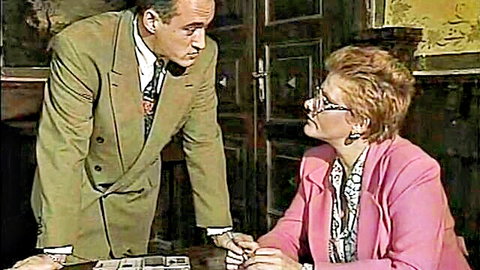 Suited man leans over desk talking to mature blonde woman in pink jacket in retro office