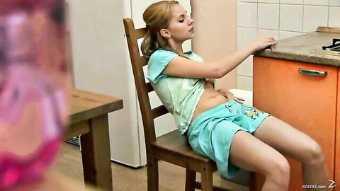 Blonde teen in green tank top and shorts sitting relaxed in kitchen solo