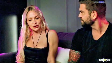 Sexy long-haired blonde in revealing black top flirts with tattooed bearded hunk on couch