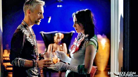 Brunette woman in green top and gloves flirts intimately with gray-haired man holding drinks in dramatic lit room