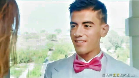 Tanned young hunk in pink bowtie suit smiles at sexy long-haired woman by window