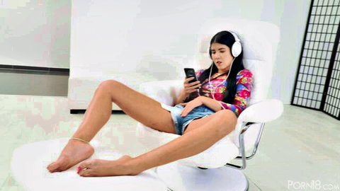 Skinny brunette teen spreading legs wide in tight denim shorts while using phone with headphones