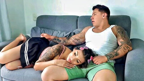 Tattooed Latina babe lies on muscular guy's lap, skirt hiked up thighs, intimate couch cuddle.