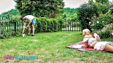 Blonde teen Novella Night lounging on blanket in white outfit, man nearby in outdoor garden