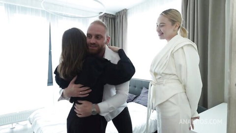 Blonde and brunette women smilingly hug pale muscular man in hotel bedroom