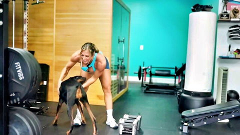 Fit blonde woman in sports bra training shoulders with Doberman dog in gym