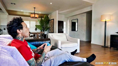 Tattooed guy in red shirt and sweatpants playing Nintendo Switch on couch in modern living room