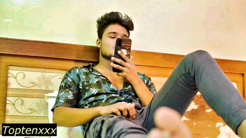 Handsome South Asian guy lounging on bed holding phone with open shirt and lowered pants teasing viewers