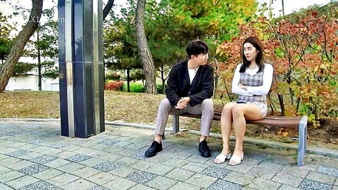 East Asian couple sitting flirtatiously on park bench, her in short plaid skirt and white top, fully clothed teasing vibe