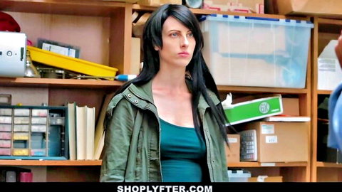 Black-haired shoplifter babe in green jacket and tank top stands in store stockroom with serious look
