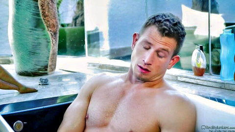 Muscular man relaxing shirtless in bathtub with woman's bare legs teasing nearby