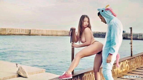 Nude skinny babe with small tits bends over pier pole as unicorn onesie guy stands behind outdoors