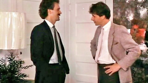 Two suited men with 80s hair converse in elegant room near lamp vintage style