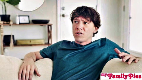 Shocked curly-haired guy in teal shirt sits surprised on couch in taboo stepsister scene