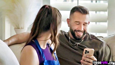 Brunette in blue apron cuddles smiling bearded man holding taboopure phone on couch