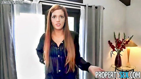 Surprised redhead babe with long hair in sheer black blouse standing by window in bedroom
