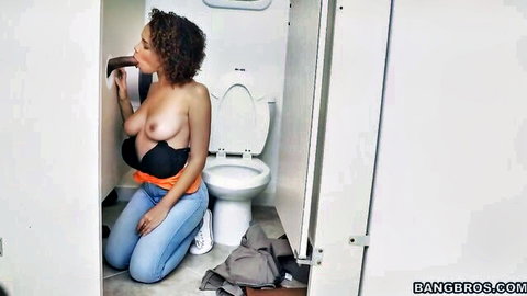 Curly haired ebony babe kneels topless in bathroom sucking thick black cock through glory hole