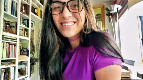 Cute Asian teen in glasses and choker smiling seductively in purple top on bookshelf