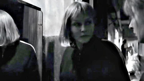 Two blonde bob-haired women in dark jackets talking intently in dimly lit kitchen scene