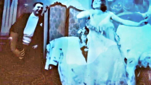 Vintage retro woman in sheer flowing dress dancing arms raised as suited man watches eagerly