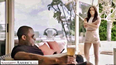 Petite brunette in tiny white shorts and crop top stands arms crossed over black stud holding beer on patio