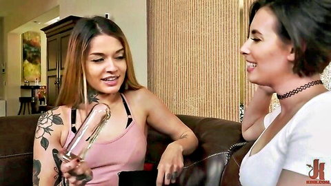 Tattooed brunette holds huge glass dildo smiling at short-haired girlfriend on couch