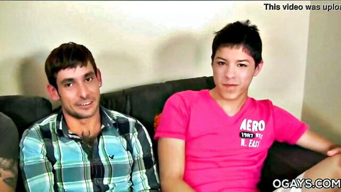 Two smiling gay twinks in plaid shirt and pink tee posing closely on couch