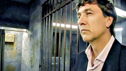 Suited man stares intently through prison bars in dimly lit cell