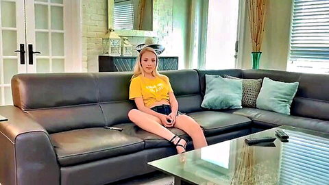 Blonde teen sister in yellow top and denim shorts sitting on couch, taboo incest vibe