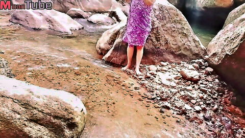 Vilashini in purple dress standing barefoot by rocky stream, outdoor tease